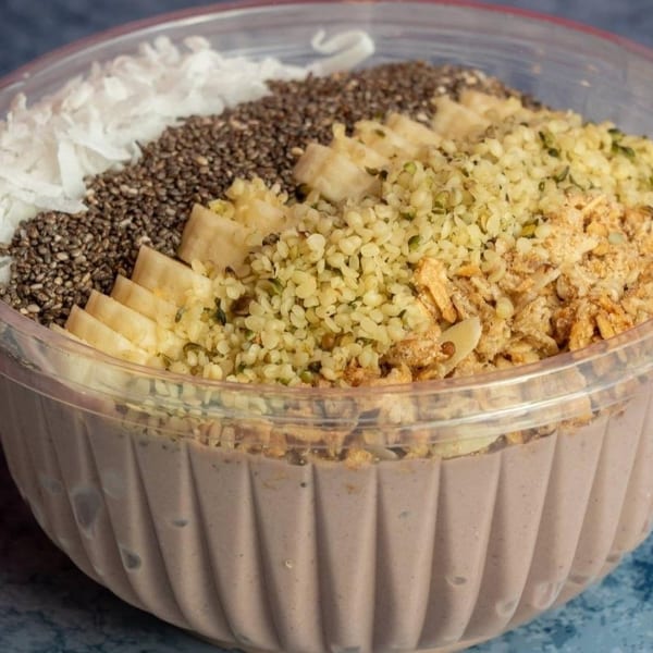 A smoothie bowl with coconut flakes, chia seeds, banana, hemp seeds, and granola in a clear plastic container.