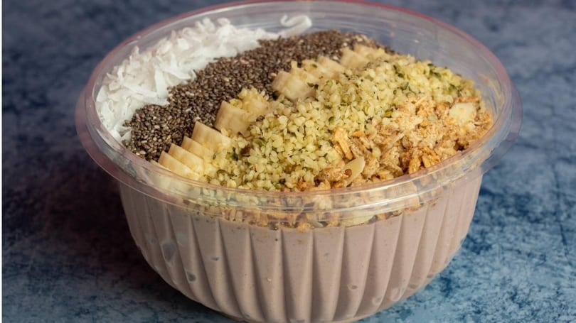 A smoothie bowl with coconut flakes, chia seeds, banana, hemp seeds, and granola in a clear plastic container.