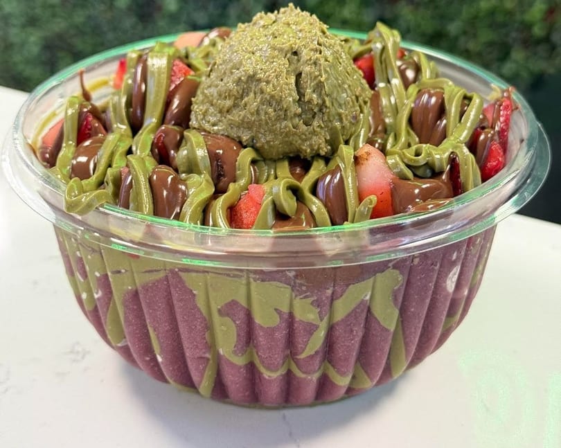 A vibrant acai bowl topped with strawberries, chocolate drizzle, and a scoop of pistachio cream.