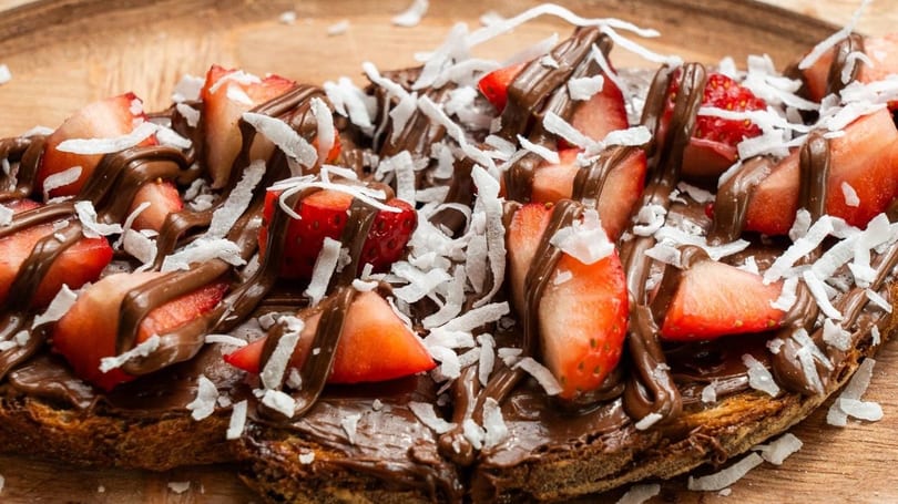 Close-up of chocolate spread on toast, topped with sliced strawberries and shredded coconut.