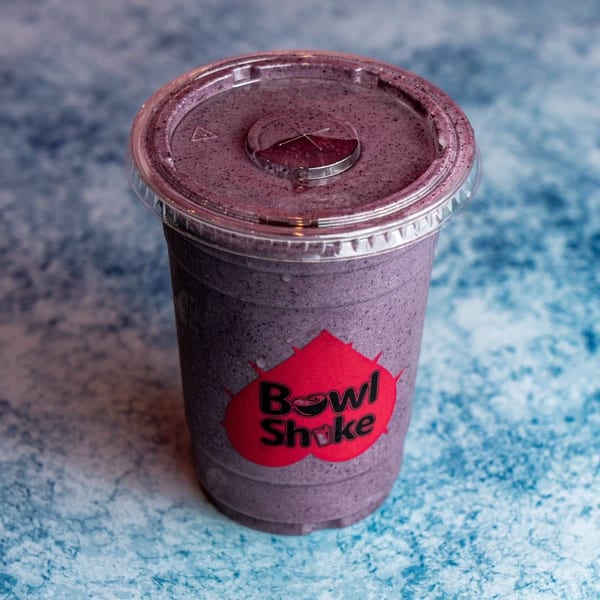 A purple Bowl Shake smoothie in a clear plastic cup with a red logo on a blue textured surface.