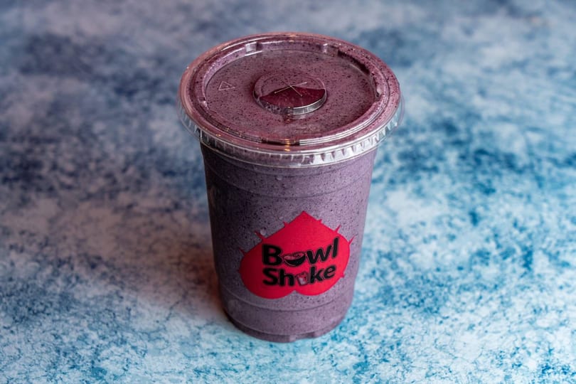 A purple Bowl Shake smoothie in a clear plastic cup with a red logo on a blue textured surface.