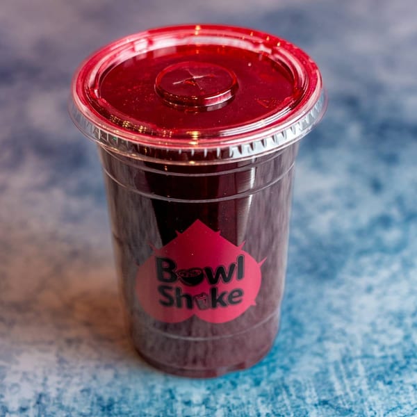 A clear plastic cup of dark red Bowl Shake smoothie with a red lid on a blue textured surface.