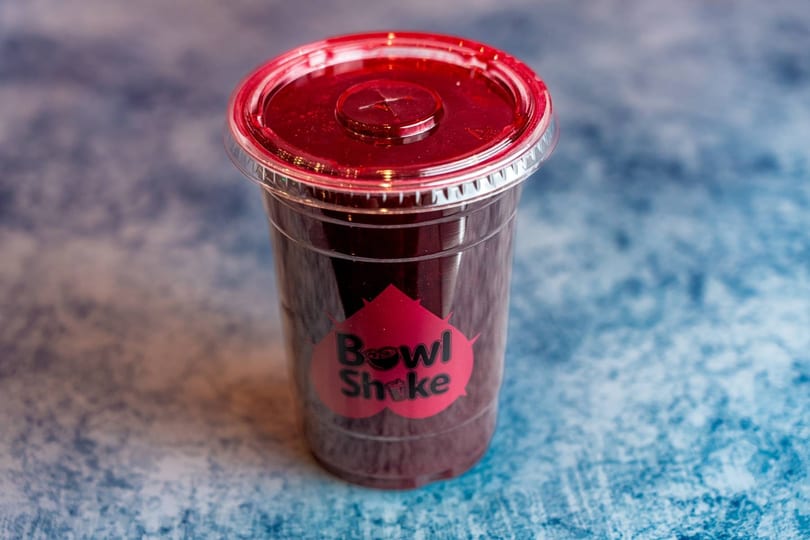 A clear plastic cup of dark red Bowl Shake smoothie with a red lid on a blue textured surface.
